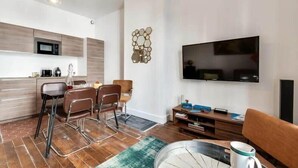 Una sala moderna con televisor de pantalla plana, un comedor con sillas, una cocina con microondas y un elemento decorativo en la pared.