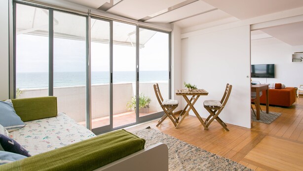 A modern living room with a large sliding glass door leading to a balcony with a view of the ocean. There is a bed with green bedding, a small table with chairs, and a TV mounted on the wall. - Sunset Bliss - Breathtaking Beach Apartment (Almada)