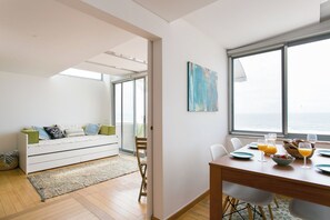 A modern living room with a sofa, a dining table set for two, and a large window with a view of the sea. - Sunset Bliss - A Breathtaking Beachfront Apartment (Almada)