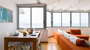 A modern living space with a dining area, a sofa, and large windows offering a view of the sea. - Sunset Bliss - Breathtaking Beach Apartment (Almada)