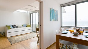 A modern living room with a sofa, dining table, and chairs. There is a large window with a view of the ocean. - Sunset Bliss - Breathtaking Beach Apartment (Almada)