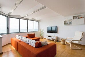 A modern living room with a large window, a flat-screen TV, a sofa, a chair, and a small cabinet. - Sunset Bliss - A Breathtaking Beachfront Apartment (Almada)