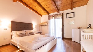 Une chambre avec un grand lit, un plancher en bois, un plafond avec des poutres apparentes, une table de chevet, une armoire et une fenêtre avec des rideaux.