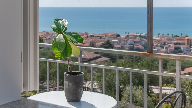 A balcony with a potted plant, a white table, and a view of the sea and town.