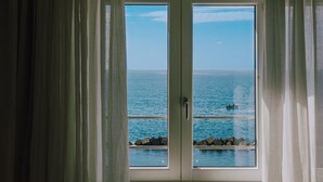 A view through a window with curtains, overlooking a clear blue sea and a rocky shoreline.