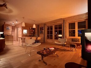 A cozy living room with a dining area, wooden walls, and large windows.