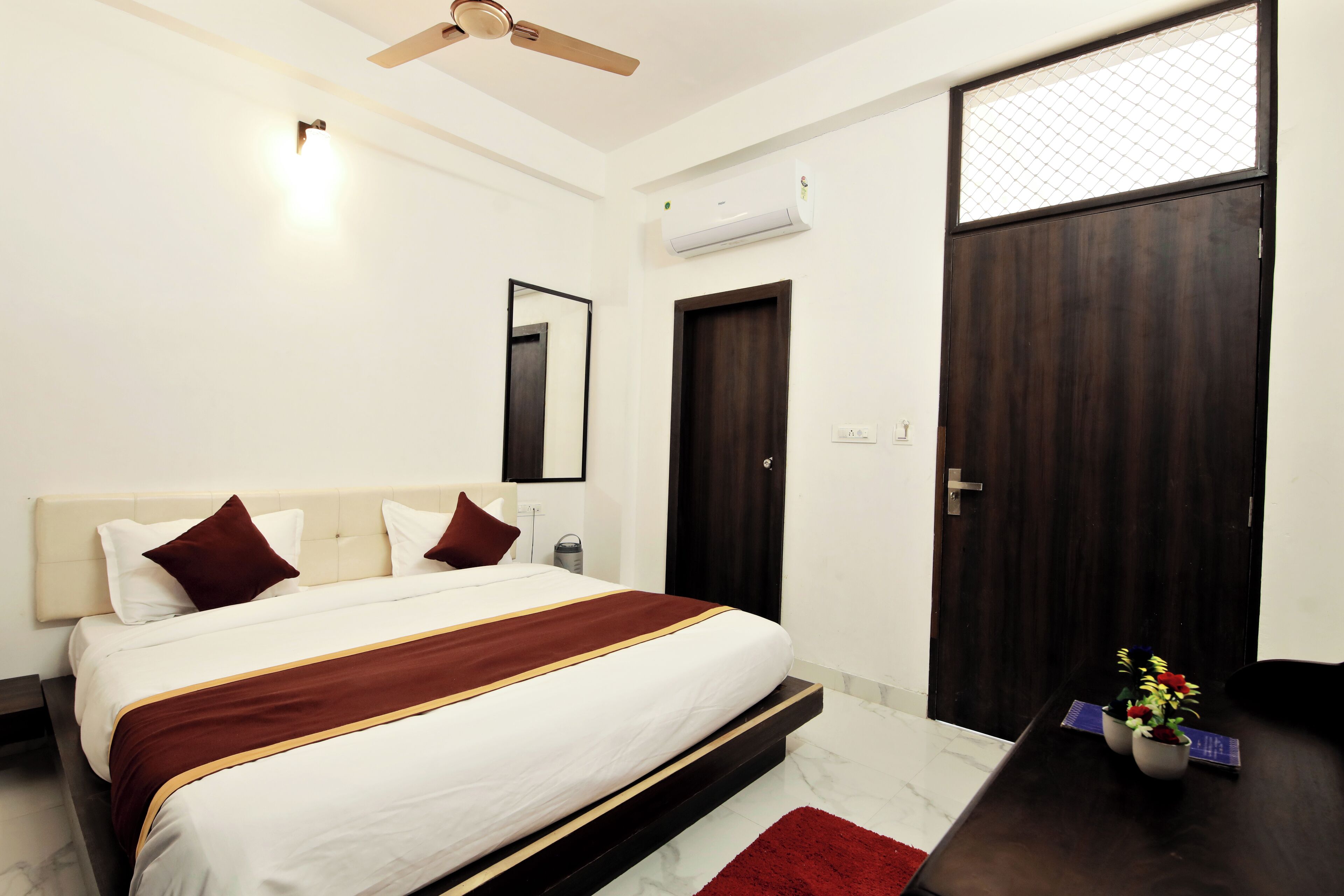 A bedroom with a bed, a ceiling fan, an air conditioning unit, a dark wooden door, and a small table with a plant.