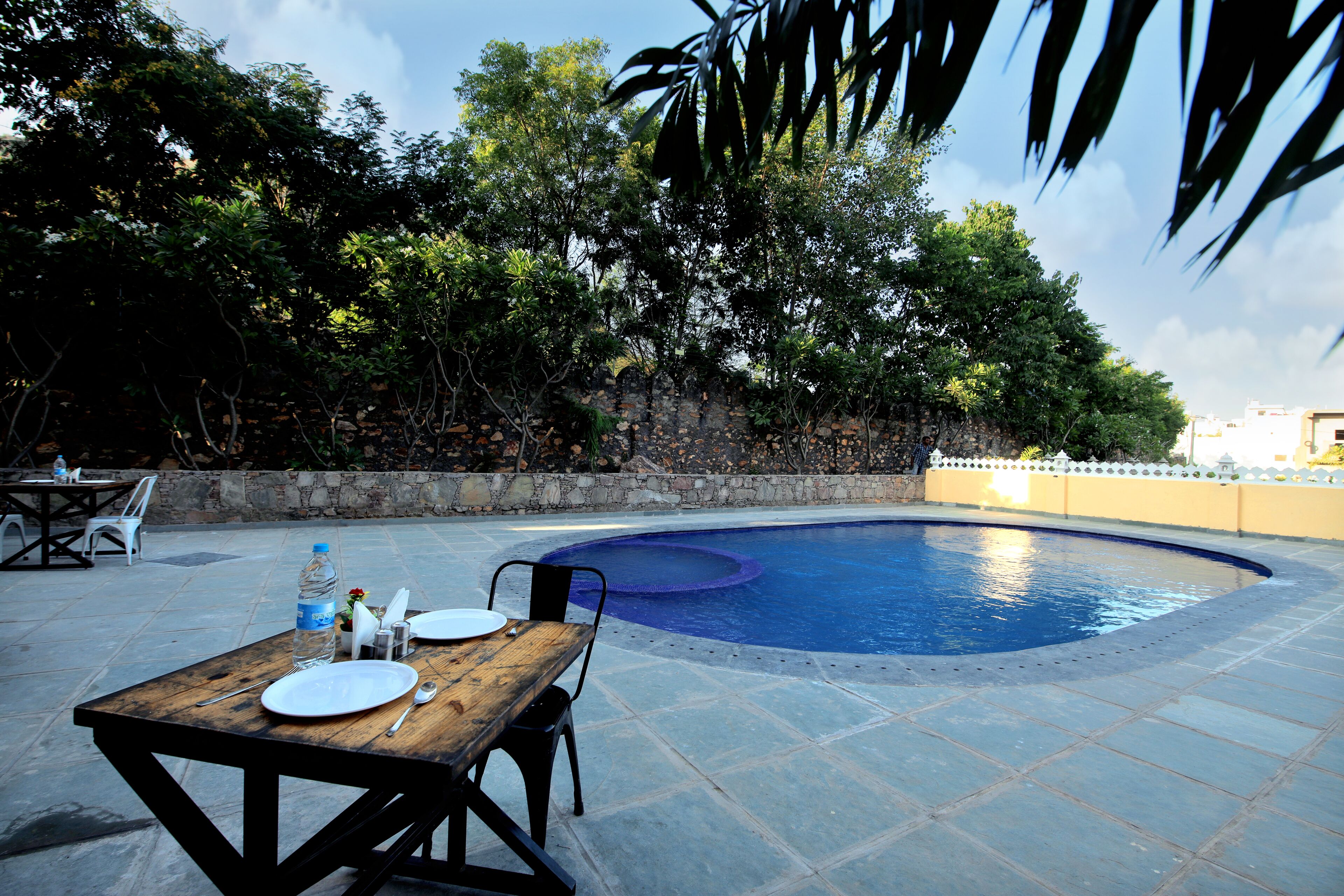 Zona de piscina al aire libre con una mesa de madera preparada para comer, rodeada de muros de piedra y abundante vegetación.