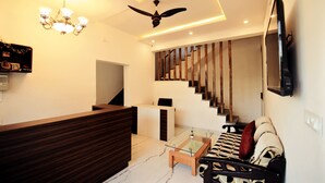 A modern living room with a wooden reception desk, a glass coffee table, a sofa with patterned cushions, a ceiling fan, and a mounted TV. - The Kanchangarh Resort (Udaipur)