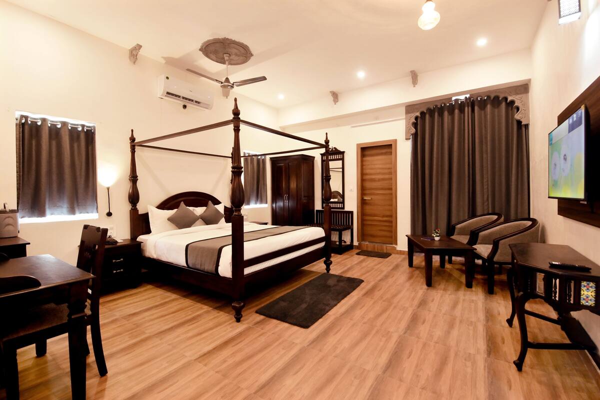 A spacious bedroom with a four-poster bed, a ceiling fan, a flat-screen TV, and a seating area with a table and chairs.