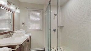 A bathroom with a glass shower enclosure, a sink with a mirror, and a window with blinds.