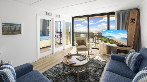 Un soggiorno moderno con una grande TV, posti a sedere confortevoli, vista sull'oceano e un balcone con terrazza.