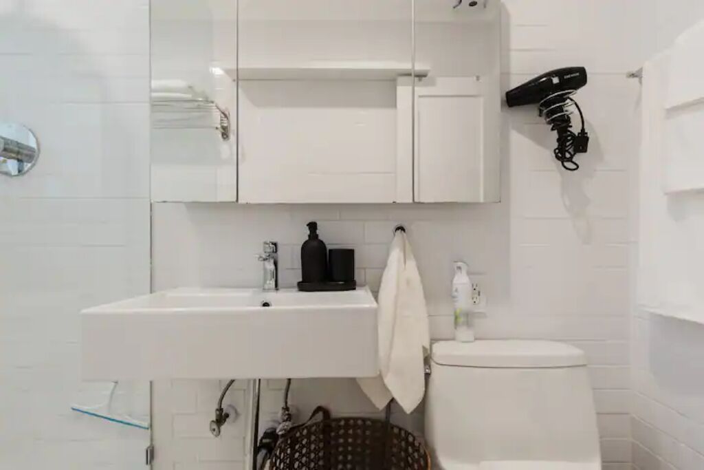 Een badkamer met een witte wastafel, spiegel en toilet. Er is een föhn aan de muur bevestigd en een mandje onder de wastafel.