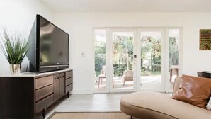 A living room with a flat-screen TV on a dark wooden cabinet, a beige sofa, and a large sliding glass door leading to an outdoor patio area. - Modern  Relaxing Zen ~ 5 ★  New pool, HotTub, Magic Garden!  (Wilton Manors)