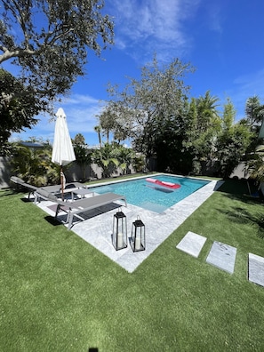 Une piscine avec un parasol blanc, des chaises longues et deux lanternes, le tout installé sur une pelouse, sous un ciel bleu dégagé.