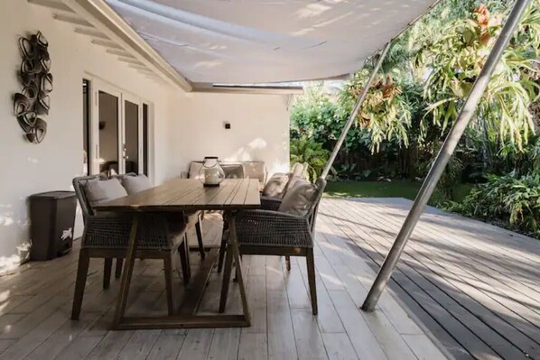 Une terrasse extérieure avec une table et des chaises en bois, entourée d’une végétation luxuriante et surmontée d’un auvent blanc.