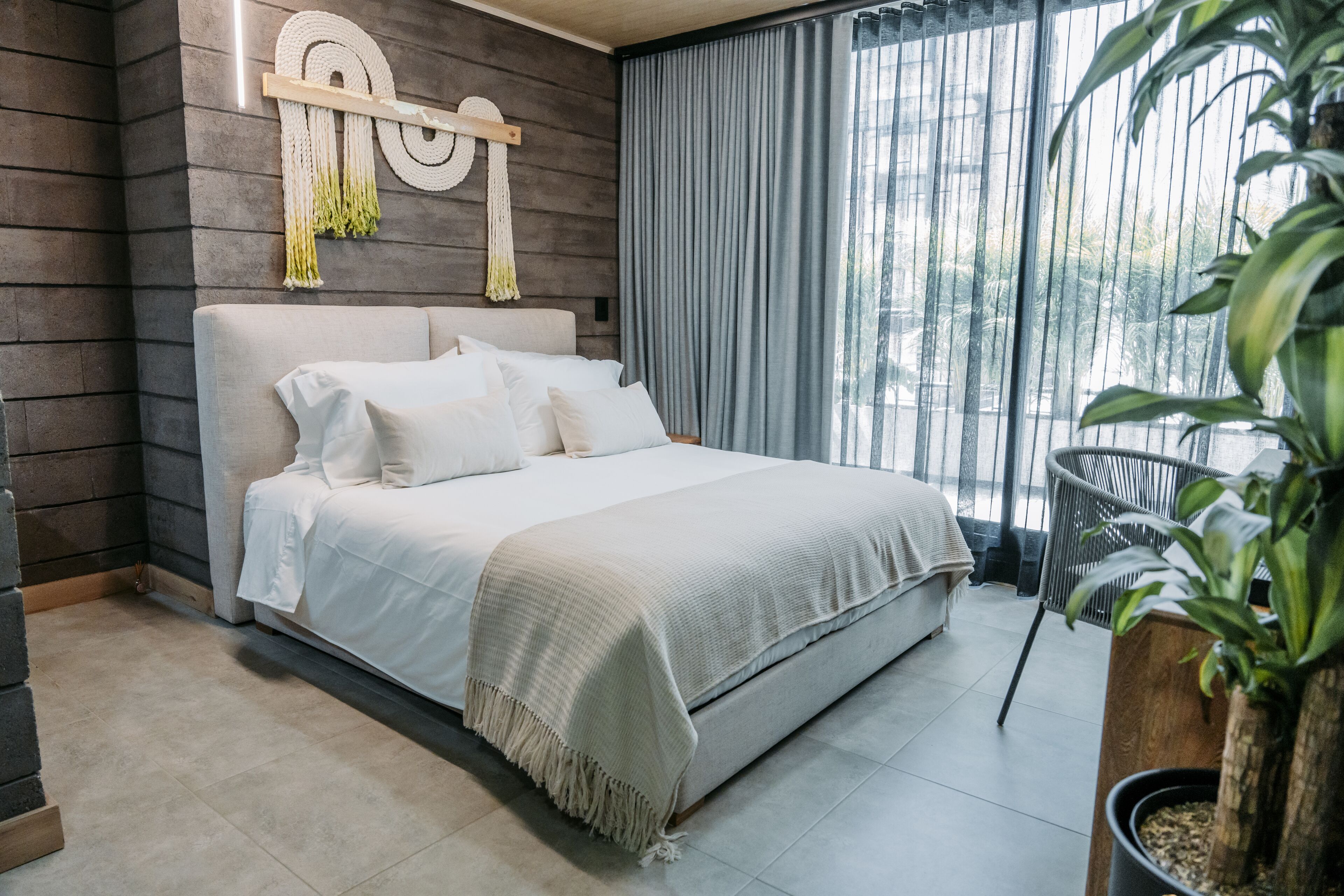 Un dormitorio moderno con una cama grande, un cabecero de madera, un colgador de macramé en la pared y vistas a zonas verdes a través de una ventana.