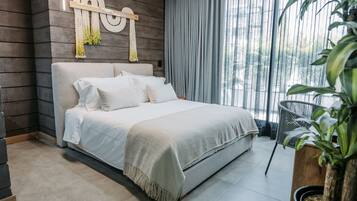 A modern bedroom with a large bed, a wooden headboard, a macrame wall hanging, and a view of greenery through a window.