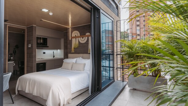 Quarto moderno com cama grande, roupeiro embutido, espaço de cozinha e varanda com plantas.