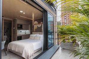 A modern bedroom with a large bed, a built-in wardrobe, a kitchen area, and a balcony with plants. - Living by Armoniko (Medellín)