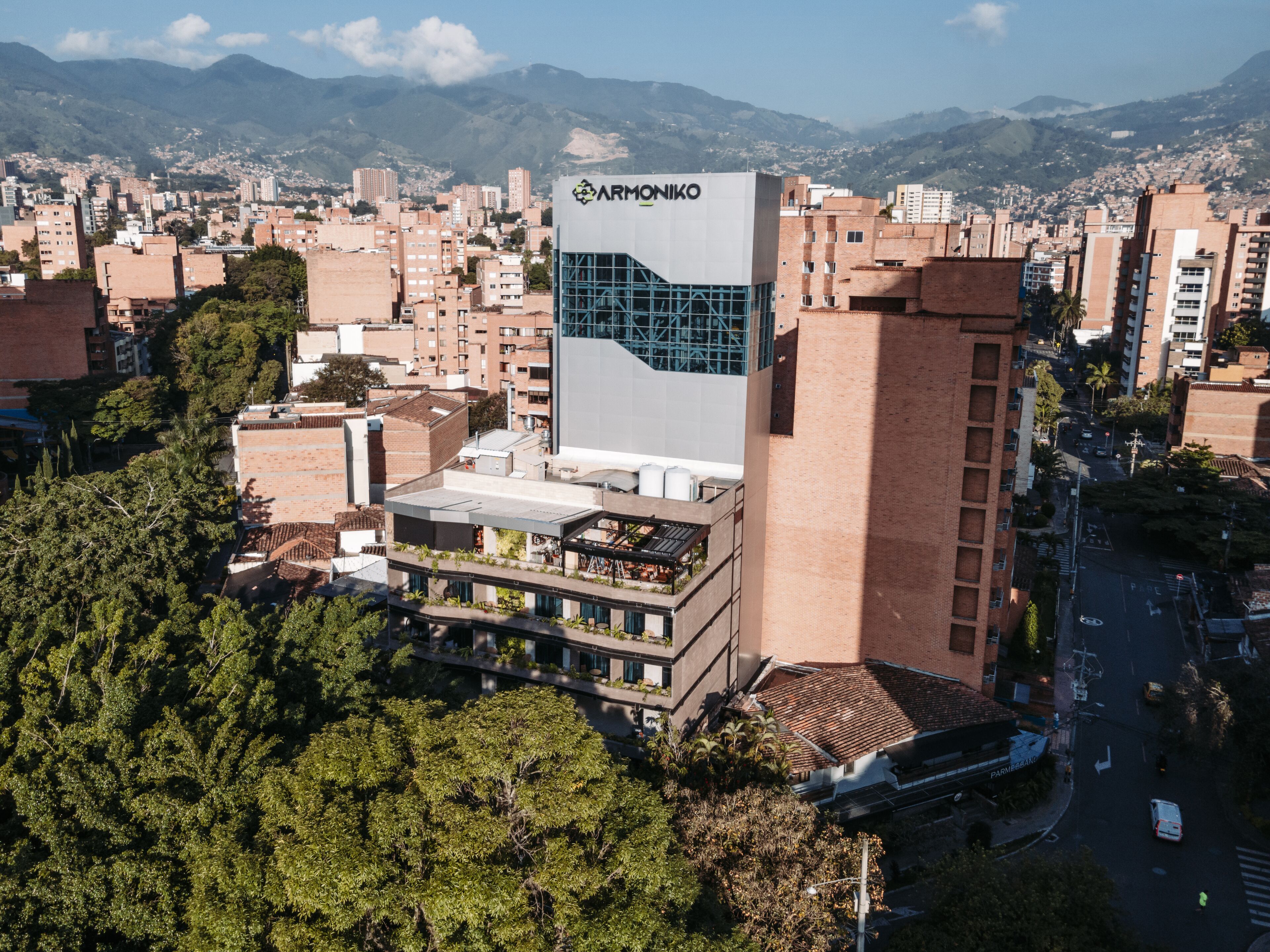 Un edificio moderno con un llamativo letrero 'ARMONIKO', rodeado de una zona urbana densa y montañas al fondo.