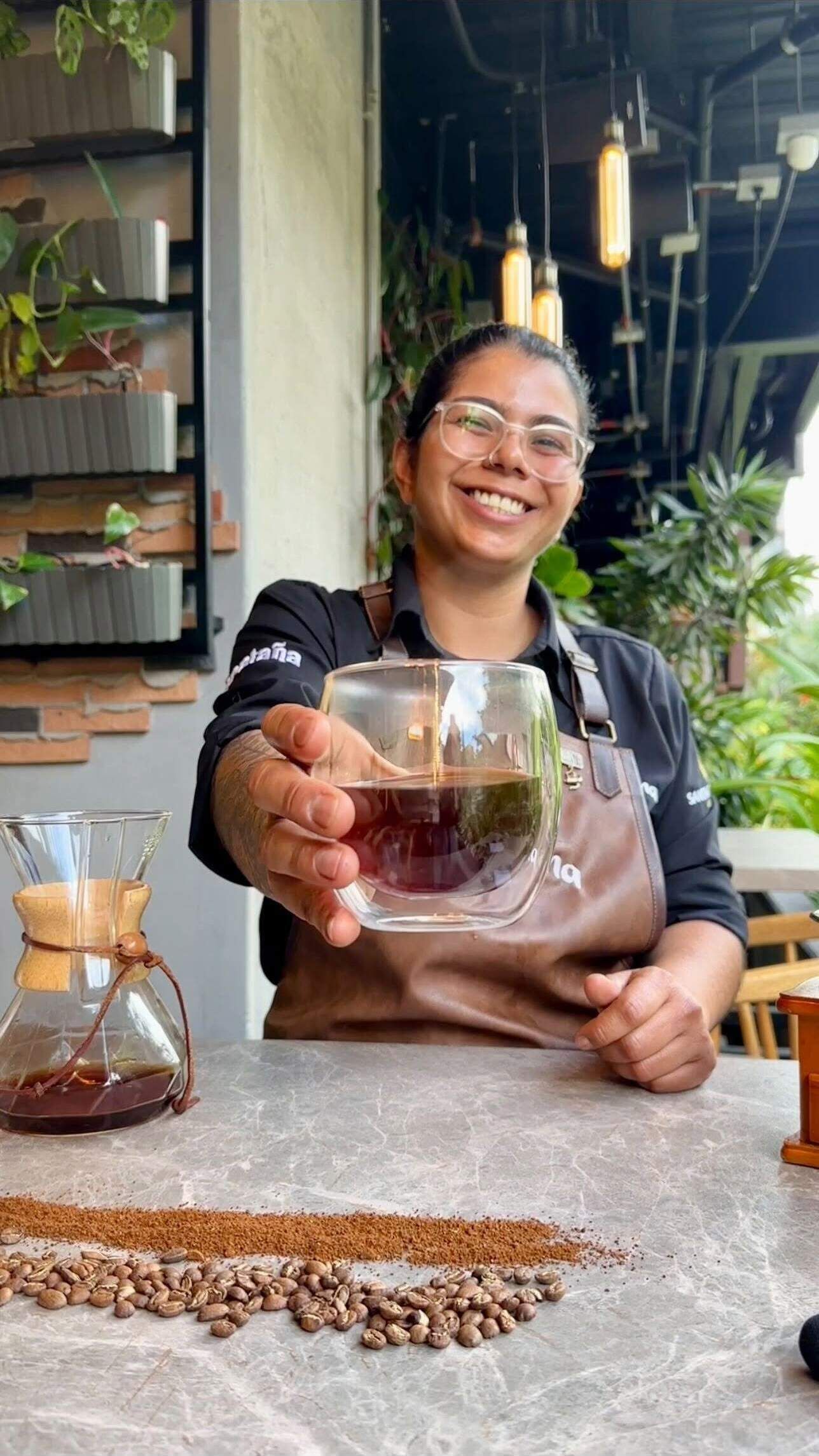 A person holding a glass of coffee at a counter with coffee beans and a coffee grinder visible.