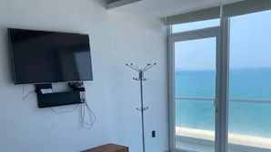 A room with a flat-screen TV mounted on the wall, a coat rack, and a large window offering a view of the sea. - Departamentos CB1 (Mazatlán)