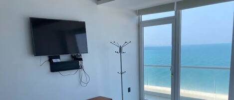 A room with a flat-screen TV mounted on the wall, a coat rack, and a large window offering a view of the sea.