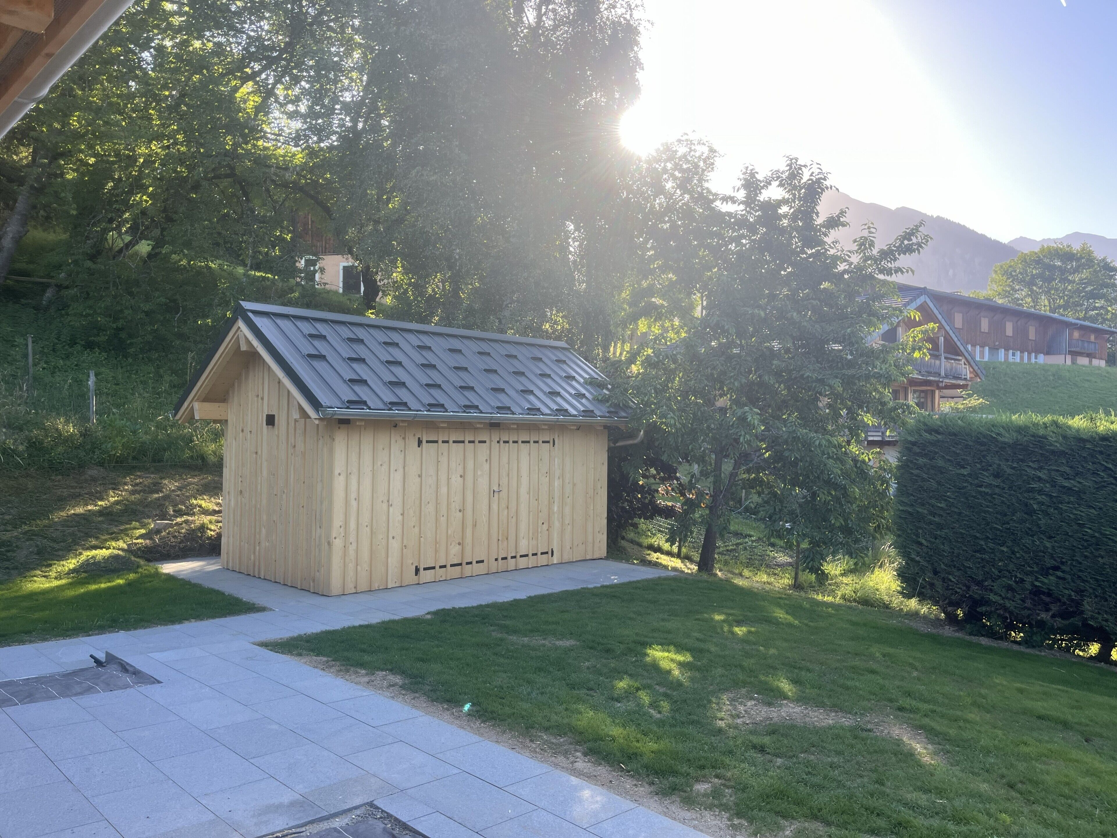 Un abri en bois avec un toit en pente, entouré d’une pelouse bien entretenue et d’arbres, avec une maison en arrière-plan.