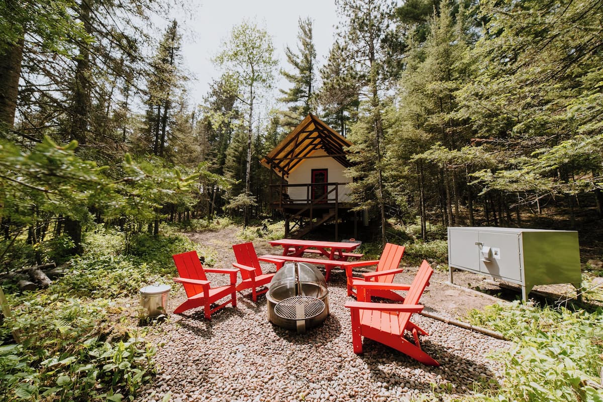 A cabin with a wooden roof, surrounded by a forest. There are red Adirondack chairs, a fire pit, and a picnic table.