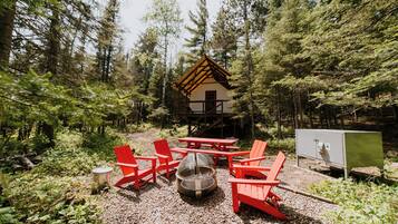 A cabin with a wooden roof, surrounded by a forest. There are red Adirondack chairs, a fire pit, and a picnic table.
