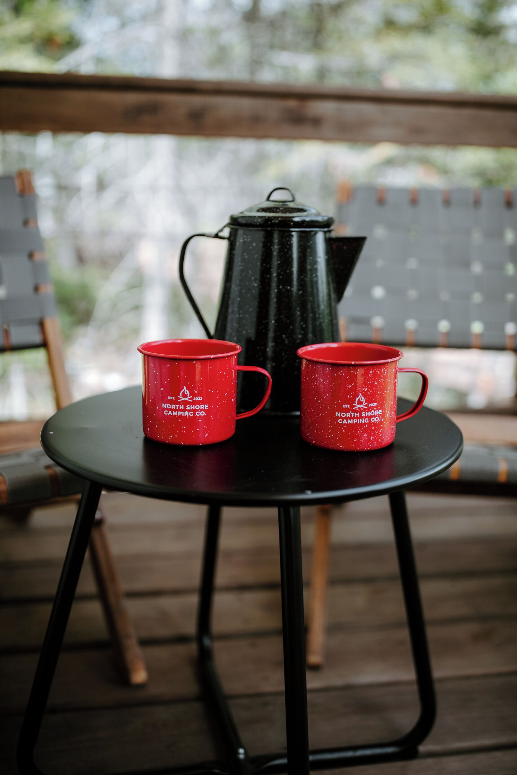A table with two red mugs and a black kettle on a wooden deck.