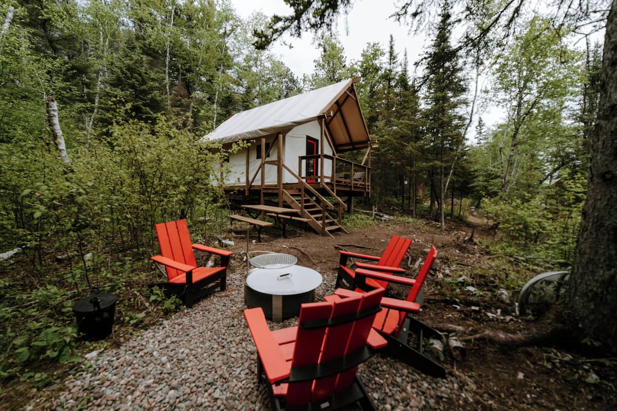 A cabin with a metal roof, surrounded by a forest. There are red chairs and a round table outside.