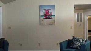 Ein Wohnzimmer mit hoher Decke, einem Strandbild, zwei blauen Sesseln und einem Korbwannen-Tisch mit Fernbedienungen und einer Zeitschrift.