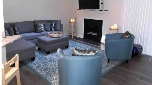 Ein Wohnzimmer mit grauem Sofa, einem blauen Sessel, einem flachbildfernseher über einem Kamin und einem gemusterten Teppich.