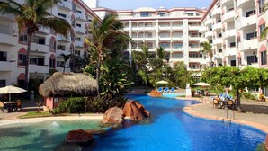 Área de piscina en un resort, con un elemento acuático central, palmeras y sillas de descanso.