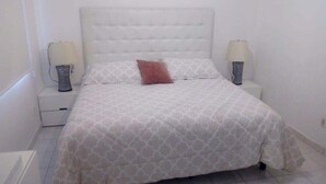 Un dormitorio con cabecera capitonada, colcha estampada, dos lámparas de noche y una mesita de noche.