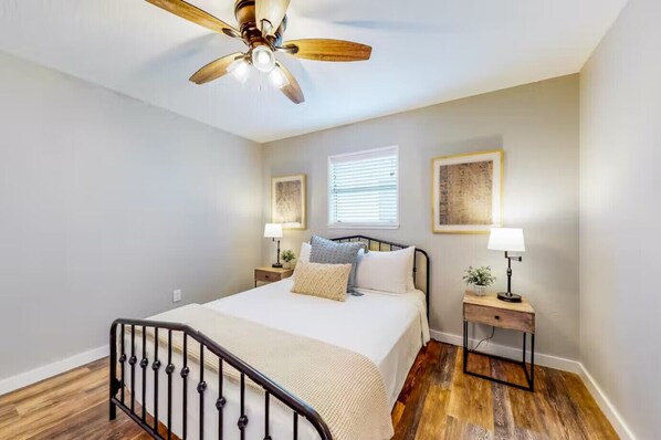 A bedroom with a bed, a ceiling fan, bedside lamps, a window with blinds, and a wooden floor.