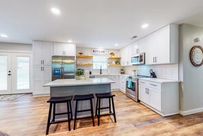 A modern kitchen with white cabinetry, a central island with stools, stainless steel appliances, and a 'KITCHEN' sign.