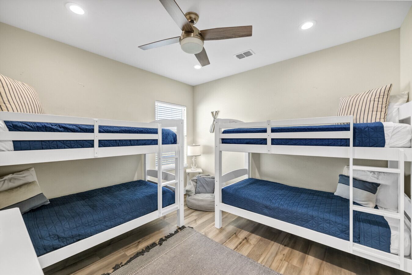 A bunk bed room with a ceiling fan, a small table with a lamp, and a window with blinds.