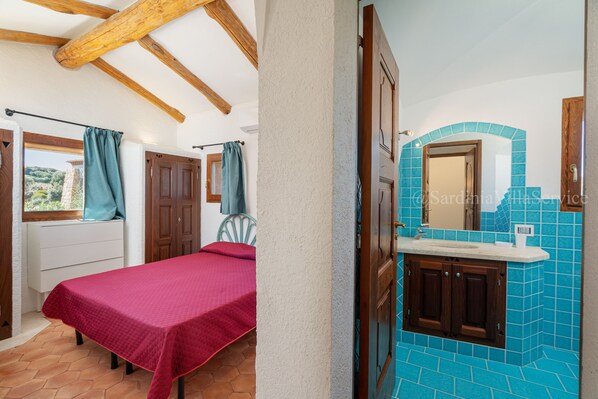 A bedroom with a bed, a window with curtains, a wooden ceiling, and a bathroom with a sink and tiled walls.