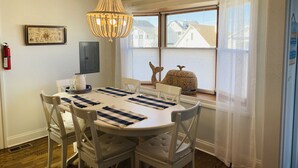 A dining area with a round table, white chairs, a chandelier, and a view of houses through a window with sheer curtains.