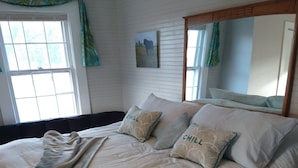 Un letto grande con una coperta e cuscini, una finestra con tende e un quadro appeso al muro.