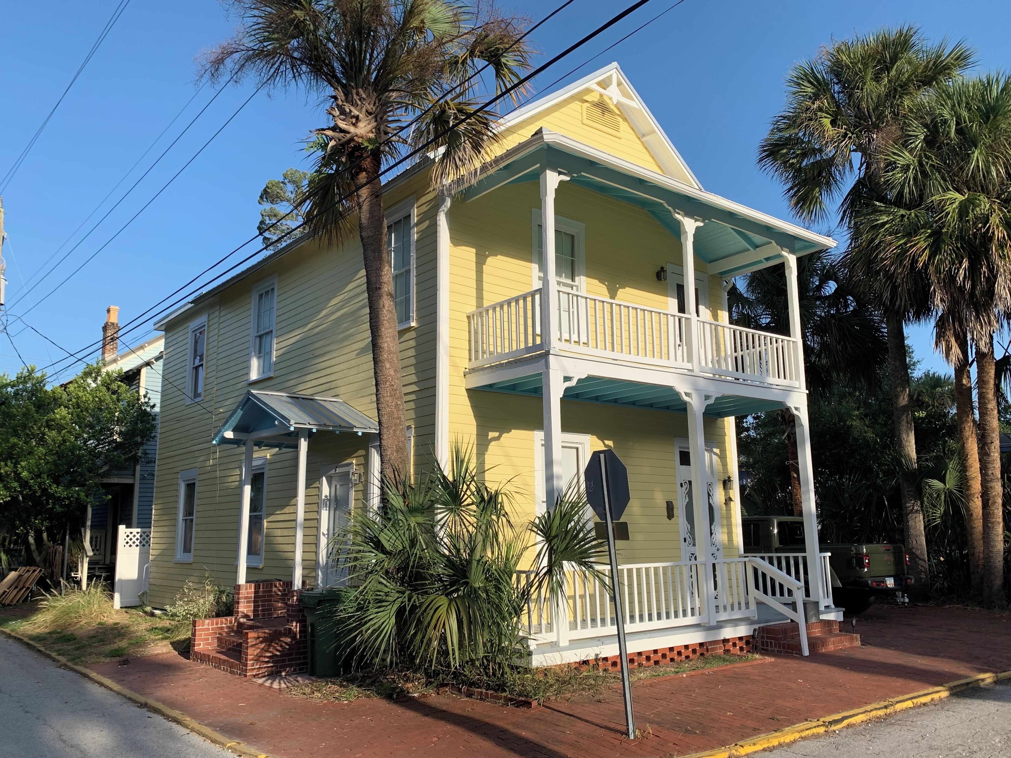 A two-story yellow house with a balcony, surrounded by palm trees and a brick foundation.