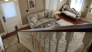 A living room with a sofa, armchair, and a glass coffee table. There is a staircase with a railing, and a door with an 'EXIT' sign. - Historic Murphy's House (St. Augustine)