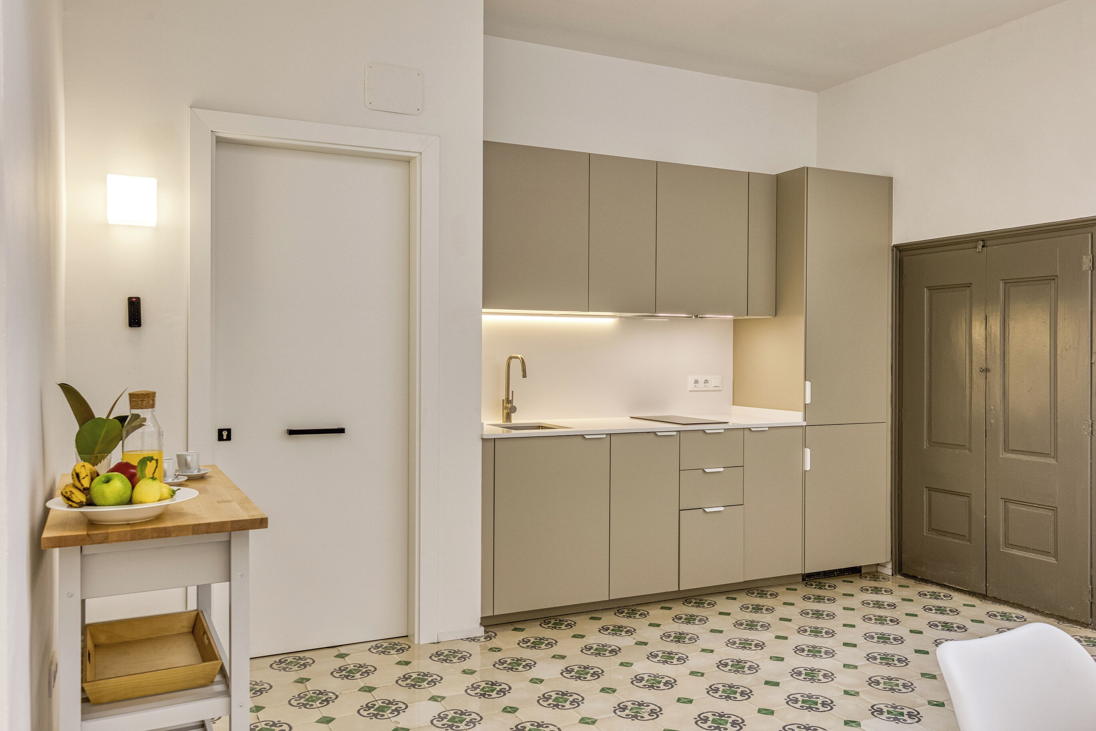 A compact kitchen with a wooden countertop, a sink, and beige cabinets. There is a small table with fruits and a bottle on it, and a patterned floor.