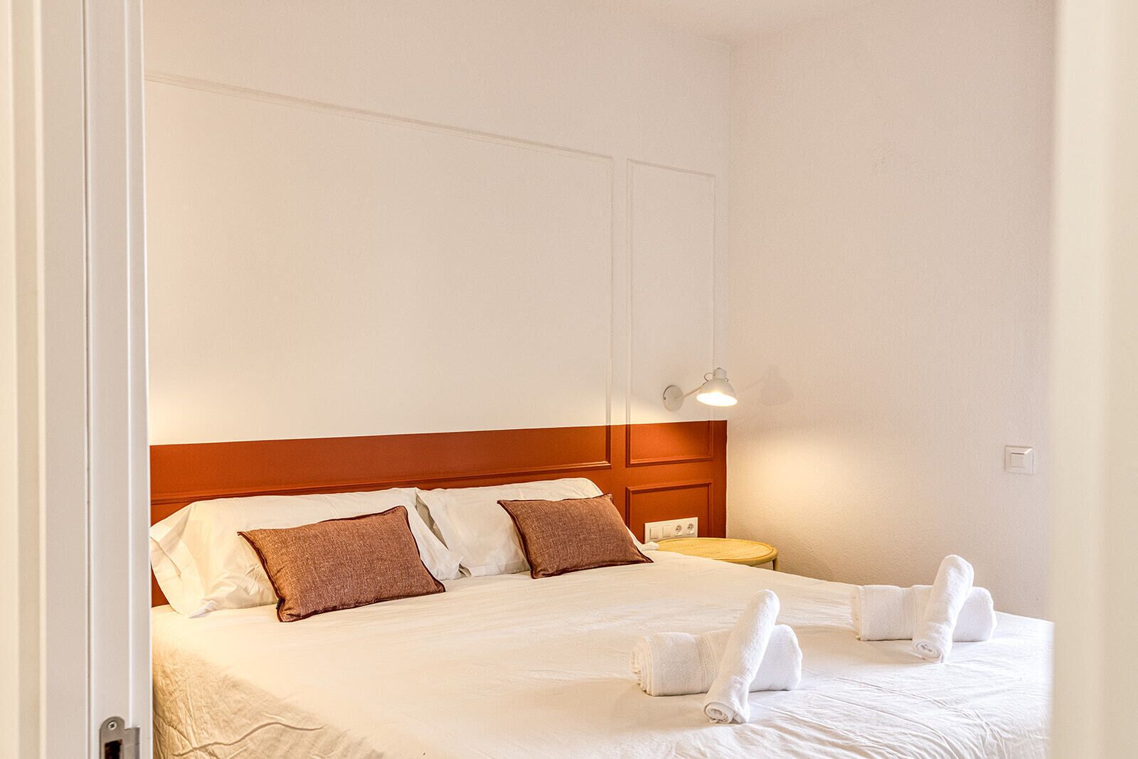 Ein ordentlich bezogenes Bett mit weißen und braunen Kissen, ein kleiner Nachttisch mit einer Lampe und ein gefaltetes Handtuch auf dem Bett.