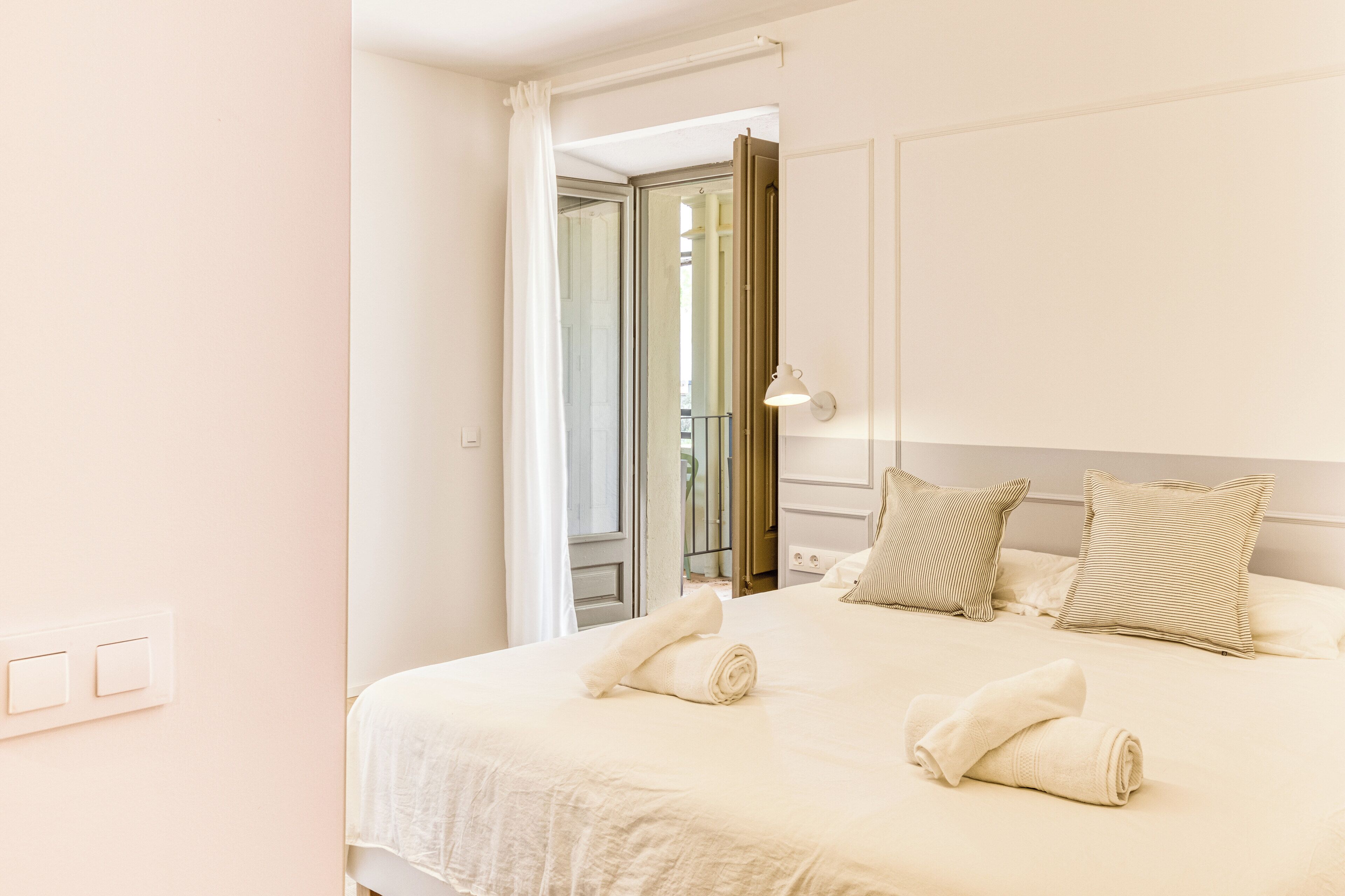Une chambre d’hôtel avec un lit bien fait, deux oreillers, une lampe de chevet et une vue sur l’extérieur, la porte étant ouverte.
