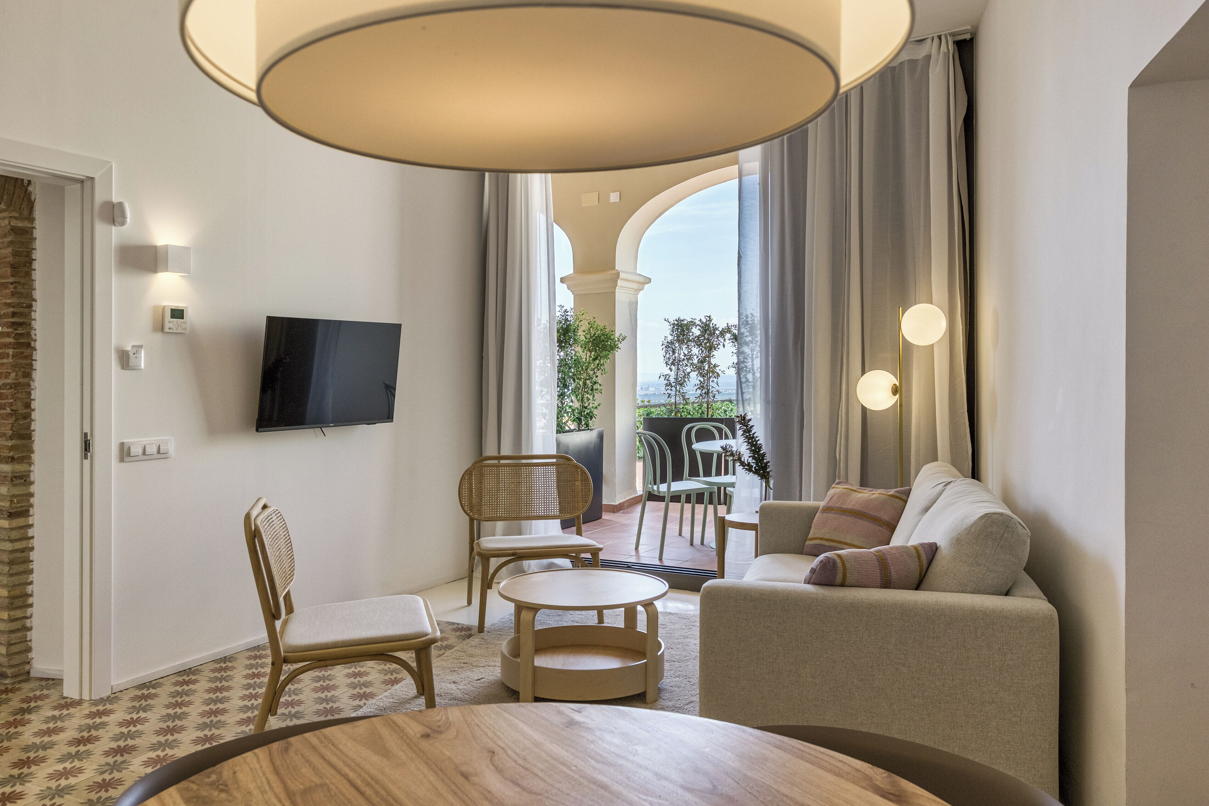 A modern hotel room with a flat-screen TV, a sofa, a dining table, and a balcony with outdoor seating.