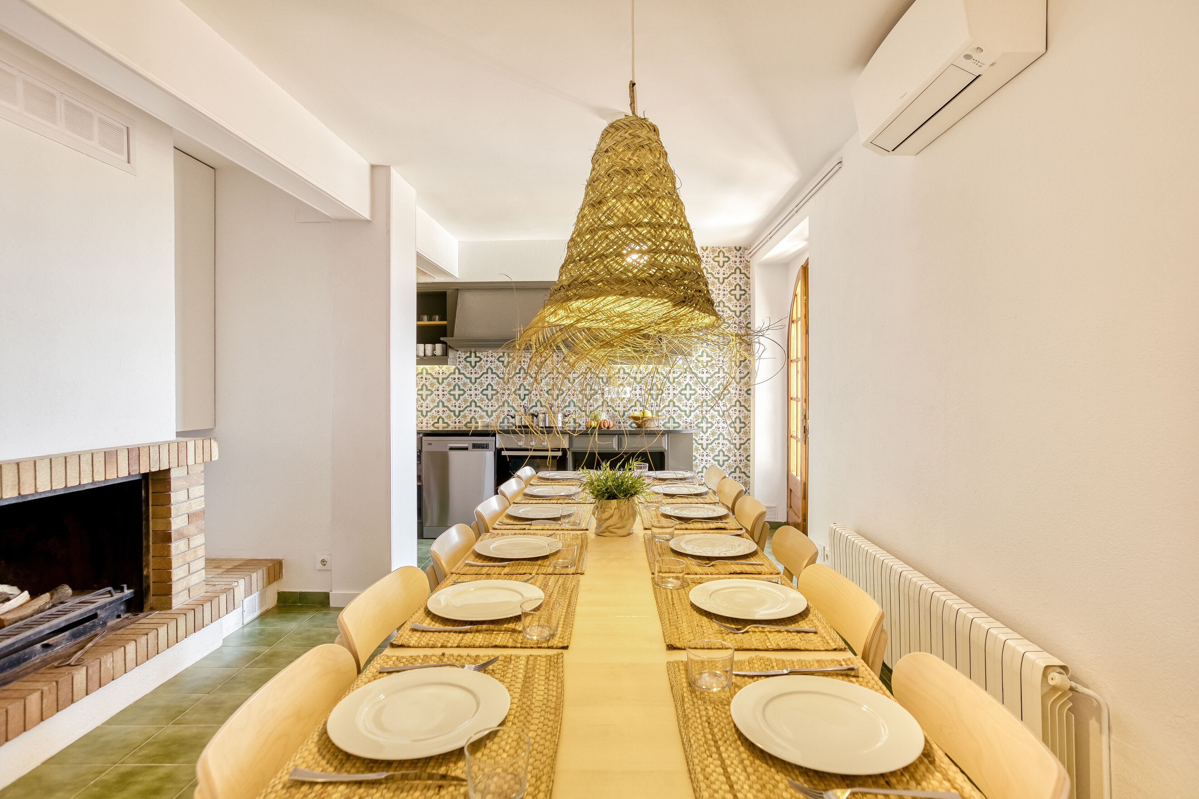 Un espace repas avec une longue table dressée pour le repas, un luminaire suspendu et une cuisine en arrière-plan.